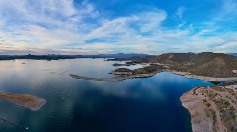 A photograph showcasing Lake Pleasant in Arizona. The image captures the lake's expansive waters surrounded by desert terrain. The shoreline features minimal vegetation, in line with the arid landscape. The clear sky overhead complements the straightforward and arid scenery.