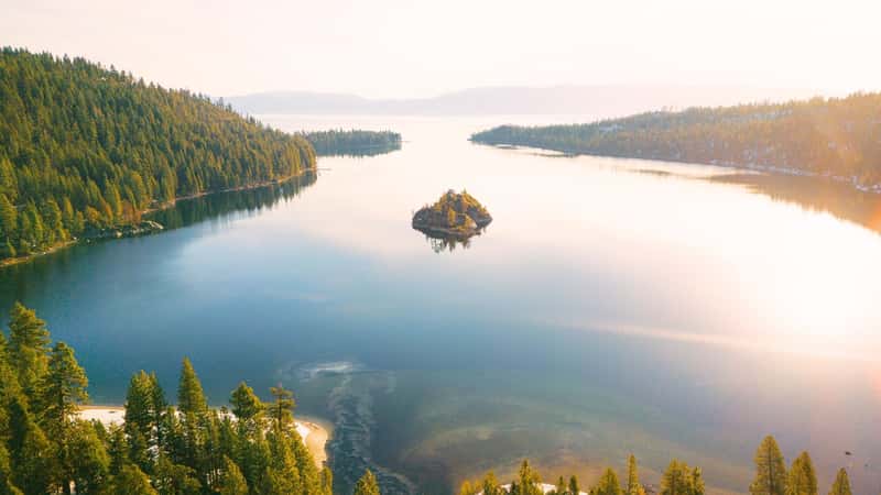 The image captures a drone shot of Emerald Bay at Lake Tahoe. The bay is characterized by its clear, emerald-green waters, surrounded by lush green forests and rocky cliffs. In the center of the bay is a small, tree-covered island, adding to the scenic beauty of the landscape.