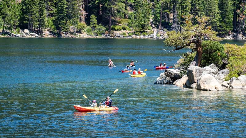 The image depicts people kayaking on South Lake Tahoe in California, USA. Six kayaks, colored in red and yellow, are visible in the frame as they navigate the serene waters. Tall pine trees and other foliage form the backdrop, highlighting the natural beauty of the lake's surroundings.