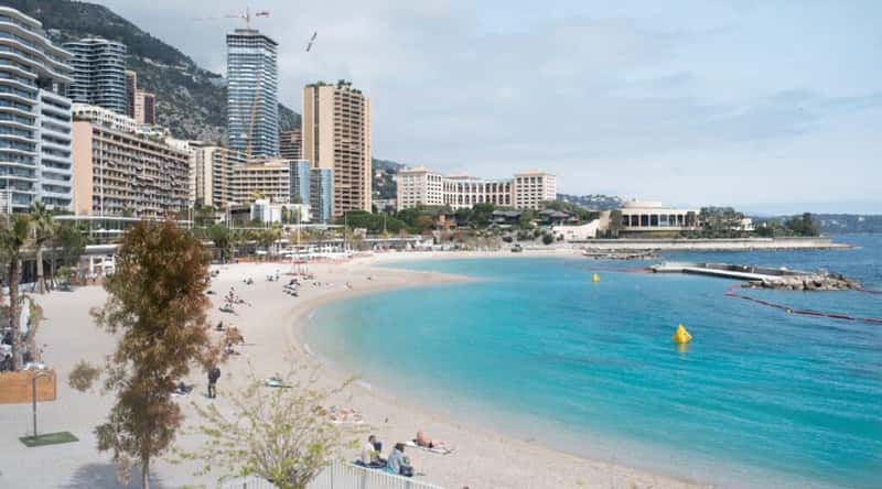 Larvotto Beach in Monaco, France. The sandy shore stretches along the azure waters of the Mediterranean Sea, creating a picturesque scene of coastal splendor. Sunbathers relax on the fine golden sand, enjoying the warm Mediterranean climate and the gentle sound of waves lapping against the shore. The turquoise water reflects the clear sky. The iconic skyline of Monaco rises in the distance.