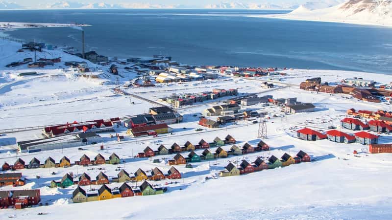 The image provides a top view of Longyearbyen, Svalbard, showcasing a snowy cityscape with houses of unique construction in various colors. The distinctive architecture adds character to the remote Arctic town. In the background, mountains and water feature prominently, highlighting the rugged natural surroundings of Svalbard.