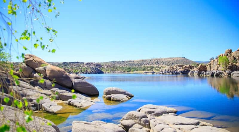 Lynx Lake in Arizona. The tranquil, crystal-clear lake kisses a stone shore adorned with medium-sized boulders. In the distance, small hills and patches of greenery complete the scenic view beneath the pure blue sky.