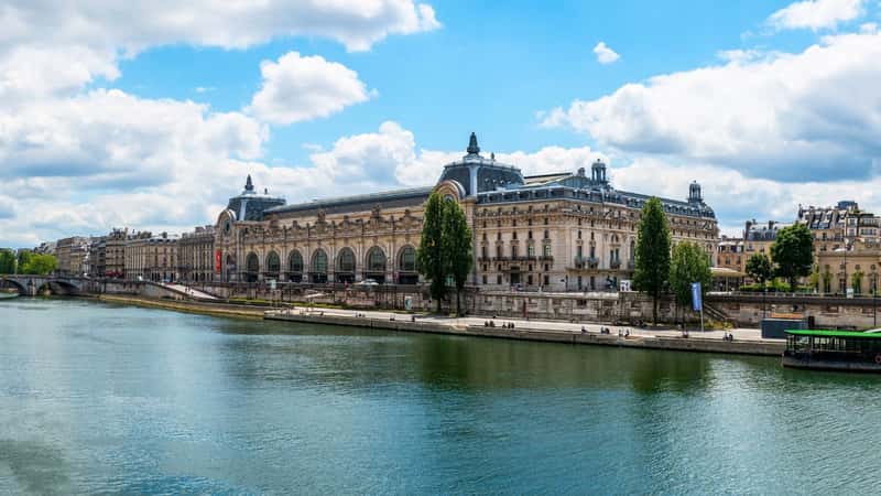 The image shows a panoramic view of the Musée d'Orsay and the Seine River in Paris, France. The museum is a prominent structure with classic French architectural features, such as ornate façades and large windows. The Seine River flows gracefully in the foreground, adding a dynamic element to the scene.