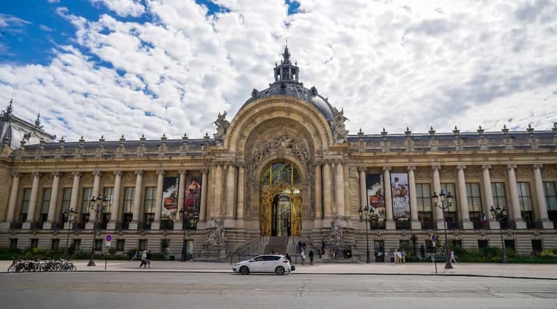 Image of the Musee du Petit Palais in Avignon, France in daylight. This museum boasts a graceful Gothic design with delicate stonework and decorative elements. Its facade features large, arched windows. The courtyard, adorned with sculptures and lush greenery. Opposite the museum there is a white car and many people are walking.