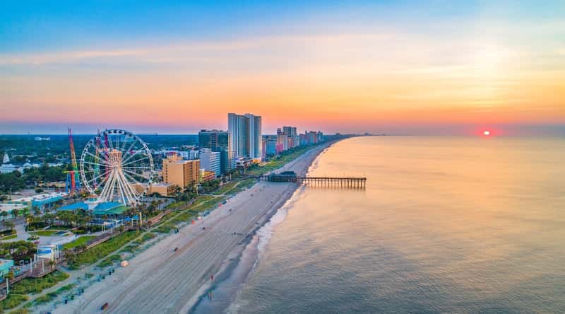 Myrtle Beach in South Carolina is a bustling vacation destination, known for its wide, sandy beaches and endless attractions, including amusement parks, shopping centers, and world-class golf courses. Top view of the red block at sunset, with white sand, a long pier, tall hotels and a beautiful Ferris wheel.