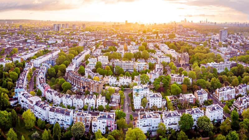 The image presents an aerial view of Notting Hill in the morning, located in London, UK. The scene features a densely populated urban area with predominantly white houses and brown roofs, characteristic of the architecture in this neighborhood. Surrounding the houses is abundant greenery, including numerous trees, enhancing the residential landscape.