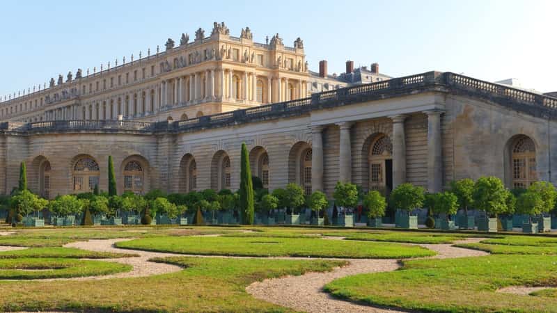 The image depicts the Orangery garden within the park of Versailles. It features neatly trimmed hedges and organized flower beds characteristic of formal French garden design. The architectural elements of the Orangery building are visible in the background.