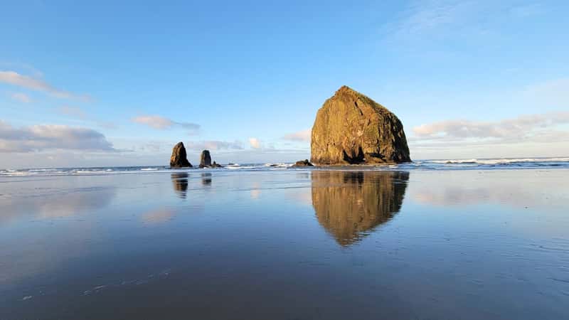 Haystack Rock in Cannon Beach, Oregon, reflected upon itself from the glassy surface it rests upon. The rock formation stands prominently against the backdrop of the beach, with its rugged texture and natural hues blending into the surroundings. The reflection creates a mirror-like effect.