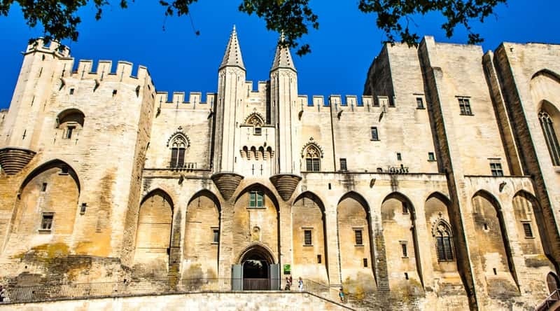 Image of the Palais des Papes in daylight. The Palais des Papes, located in Avignon, France, is an imposing medieval fortress and palace. This massive stone structure features impressive Gothic architecture, characterized by its soaring walls, pointed arches, and intricate detailing. The palace's facade showcases a harmonious blend of dark, weathered stone and ornate carvings.