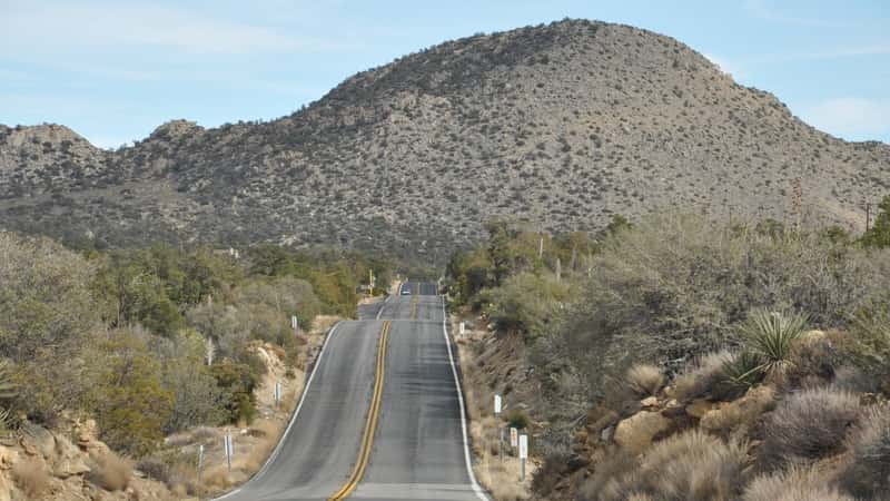 The image depicts the Pines to Palms Scenic Byway in California, showcasing a winding road bordered by tall mountainous terrain. The clear blue sky overhead complements the natural scenery.