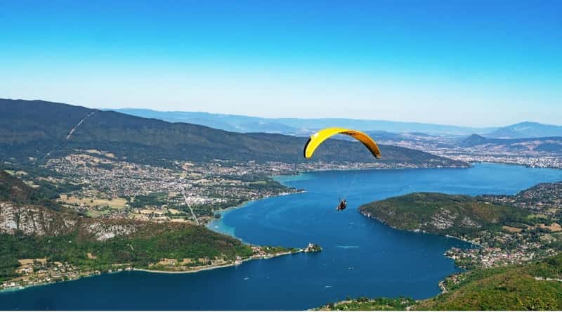 View of paraglide over Lake Annecy. The aerial view of Lake Annecy is breathtaking, offering a panoramic vista of its stunning natural beauty. Nestled within the embrace of the majestic French Alps, the lake's crystal-clear waters shimmer in various shades of blue and green. The surrounding landscape showcases quaint lakeside villages, lush forests, and the charming town of Annecy.