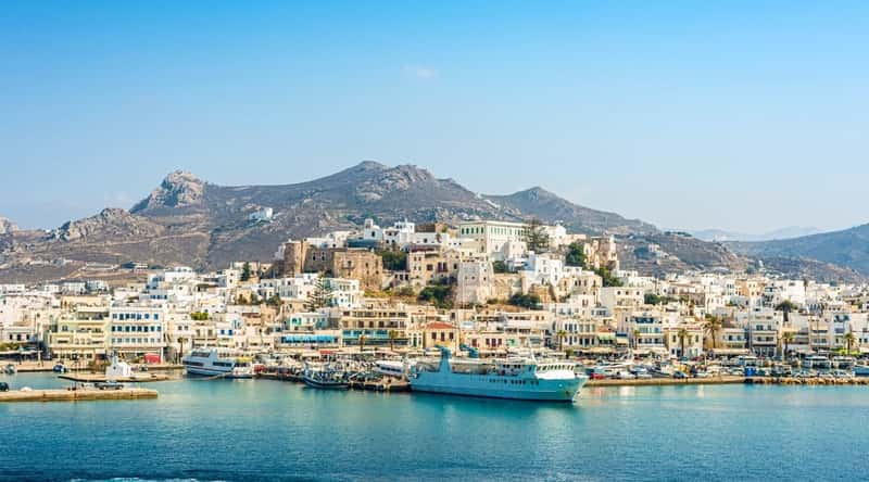 An image capturing the skyline of Paros Island Port in the central Aegean Sea, Greece. The photograph presents a view of the port area with a focus on the architectural elements and surrounding landscape. The port features a collection of buildings along the waterfront, possibly indicative of commercial and maritime activities.