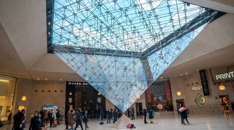 A photograph capturing the interior of the Louvre Museum, featuring people within the art gallery. In the foreground, there is an inverted glass pyramid extending from the ceiling to the floor.