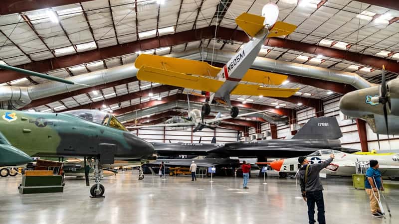 Pima Air & Space Museum, showcasing numerous planes on display. Visitors are seen inspecting the aircraft. Some planes are suspended on cables at a height. 