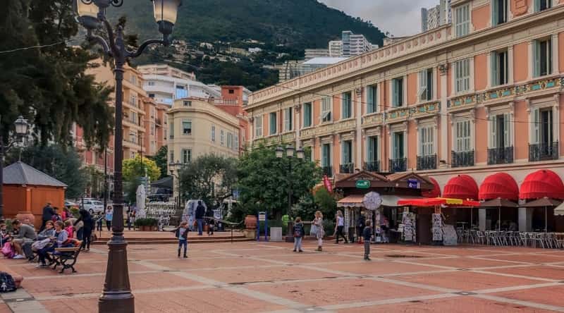 Place d'Armes in Monaco, France. The square is a bustling hub of activity, surrounded by elegant architecture and vibrant cafes. In the foreground, locals and tourists alike can be seen strolling across the square's cobblestone pavement, creating a lively and cosmopolitan atmosphere.