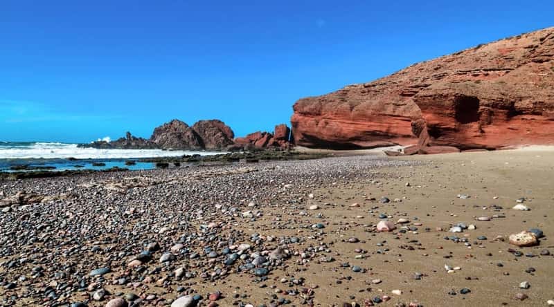 Plage de la Garrigue in Saint Tropez, France, unfolds with a shore covered in small gravel. Beyond the beach, a red-brown rocky hill emerges, complementing the pure blue sky overhead. The sea, adorned with waves.