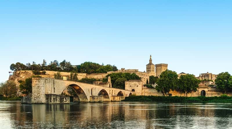 View of Pont Saint-Benezet. The Pont Saint-Benezet, also known as the Pont d'Avignon, is a picturesque and iconic bridge in Avignon, France. This historic structure has a series of graceful stone arches that extend only partway across the Rhone River. Green trees and elements of a stone bridge are reflected in the river.