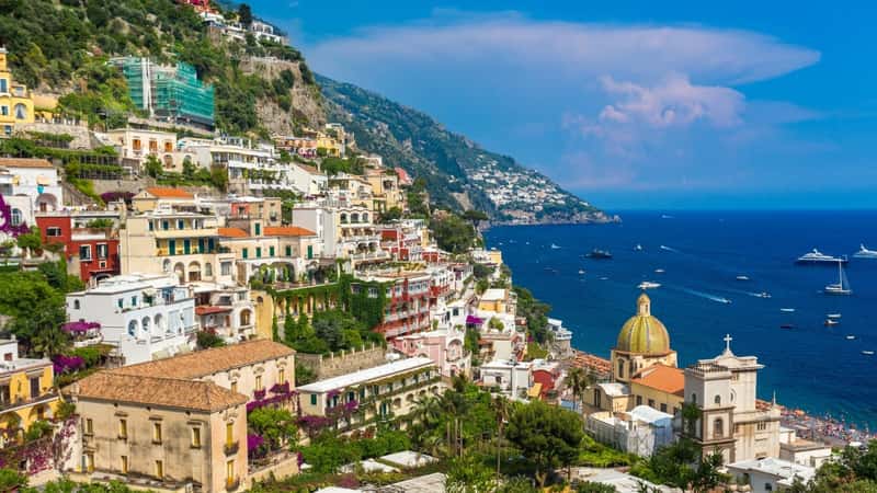 The image captures Positano, Italy, showcasing the city perched on a hillside with a variety of colorful houses. The houses exhibit different hues, creating a vibrant and picturesque cityscape. In the background, the ocean is visible, dotted with yachts and ships, adding a coastal element to the scene.