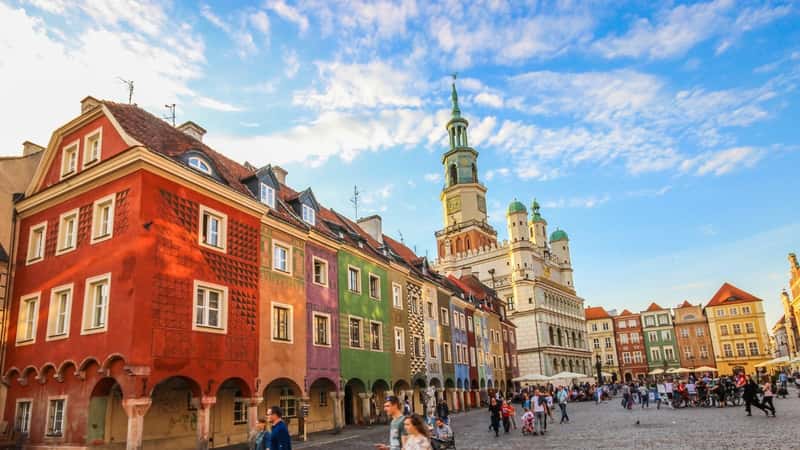 The image depicts the main square of the old town of Poznan, Poland, bustling with numerous people. Surrounding the square are colorful houses in bright hues, contributing to the vibrant atmosphere of the area. Dominating the scene is a church with a high dome, adding to the architectural diversity of the square.