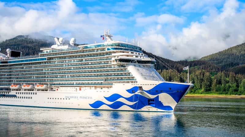 The image features the Cruise ship Discovery Princess, characterized by its white exterior with blue wave patterns on the front of the ship. In the background, forested mountains create a scenic backdrop for the ship.
