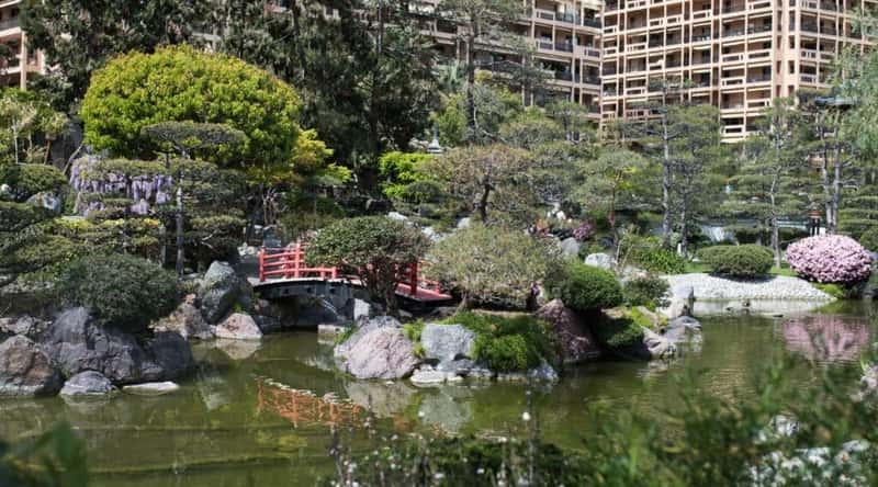 The Princess Grace Japanese Garden in Monaco is depicted in the image, highlighting intricate landscaping with meticulously maintained Japanese flora that forms a tranquil oasis. Architectural elements feature arched bridges spanning meandering streams, offering scenic views of the ponds.