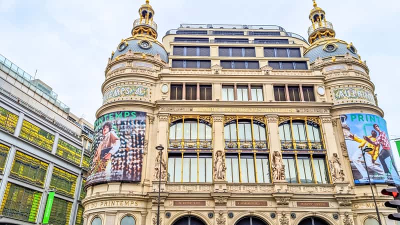 The image captures a view of the Printemps department store in Paris, France. The building features a blend of modern and classic architectural elements, with ornate details and a grand facade. The color scheme includes shades of cream, beige, and gold, typical of historic Parisian architecture.