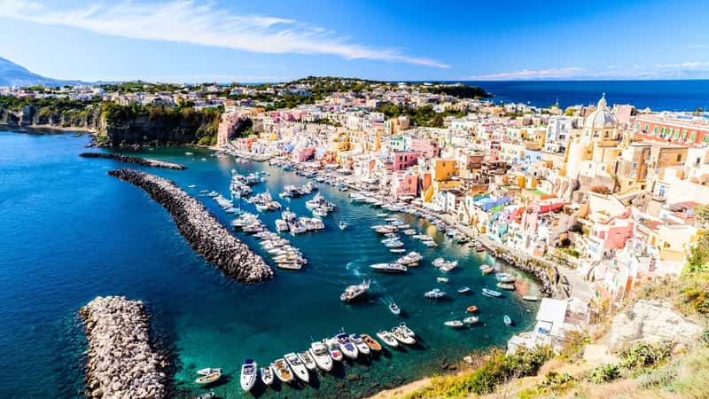 The image showcases Procida Island, Italy, known for its densely populated cityscape characterized by brightly colored houses. Along the shore, numerous yachts are moored. The colorful buildings create a picturesque contrast against the blue waters of the Mediterranean Sea.