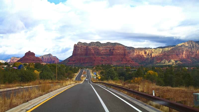 A photograph depicting a scenic drive along Red Rock Scenic Byway in Sedona, Arizona. The image showcases the picturesque landscape characterized by red rock formations and desert vegetation. 