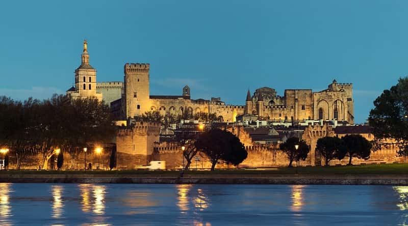View of Remparts d'Avignon in the evening illuminated by yellow lanterns. The Remparts d'Avignon, or the city walls of Avignon, has sturdy stone walls and watchtowers. The walls are punctuated by imposing gates and battlements. In front of this medieval fortifications there is a beautiful green square with trees and a wide river.