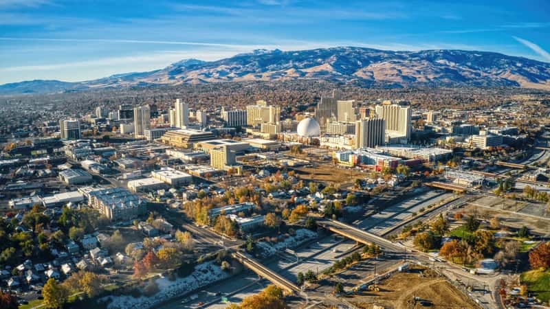 Reno, Nevada, offers a panoramic view from a drone, highlighting the city's blend of urban and natural elements. The cityscape features a mix of buildings, roads, and green spaces, indicating a balance of development and vegetation. The backdrop includes mountains, presenting a stark contrast with the city's structures.