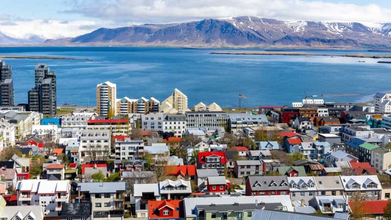 The image depicts Reykjavik, Iceland, showcasing a landscape view of the city characterized by colorful residential buildings. In the background, there is an ocean view along with the snowcapped Mount Esjan mountain range, adding a scenic backdrop to the urban setting.