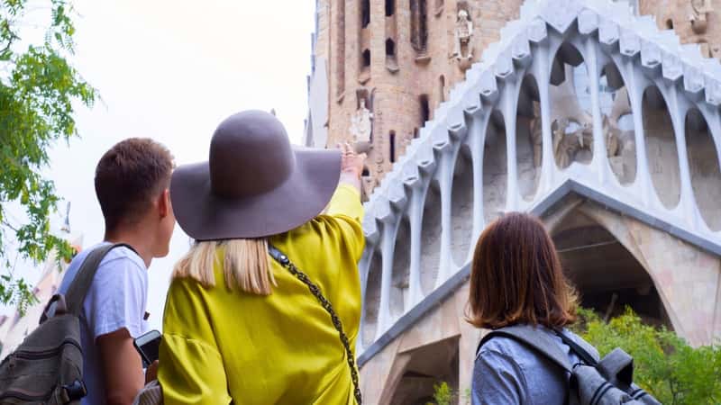 The image depicts a girl wearing a hat, showing something on the Temple Expiatori de la Sagrada Família to two men with backpacks. The scene captures the interaction between the individuals and the architectural details of the temple.
