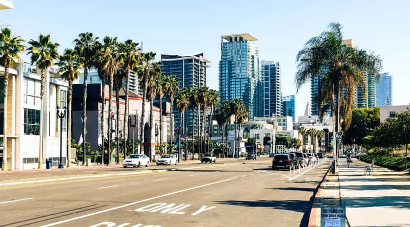 A street view in San Diego showcasing a wide road with moving traffic, including cars, in the foreground. The thoroughfare is lined with palm trees, contributing to the region's characteristic landscape. In the background, high-rise buildings form the urban skyline.