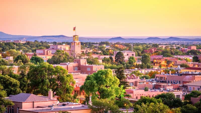Downtown skyline of Santa Fe, New Mexico, USA, captured during dusk. The scene includes numerous trees interspersed between the houses, adding a touch of greenery to the urban environment.