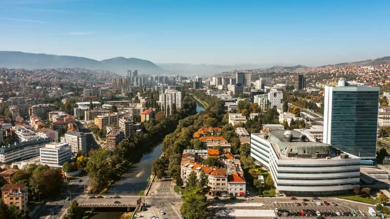 A photograph capturing the cityscape of Sarajevo. The image showcases urban architecture and a mix of buildings against a neutral sky. The landscape includes streets, pedestrian areas, and green spaces.