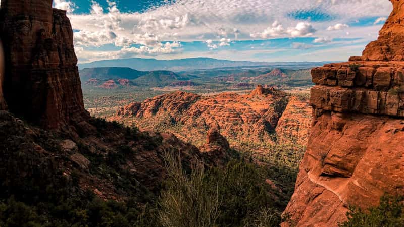 Cathedral Rock trail in Sedona, Arizona, offering a picturesque view of the red-brown rocky mountains typical of the region. The sky is adorned with white clouds.