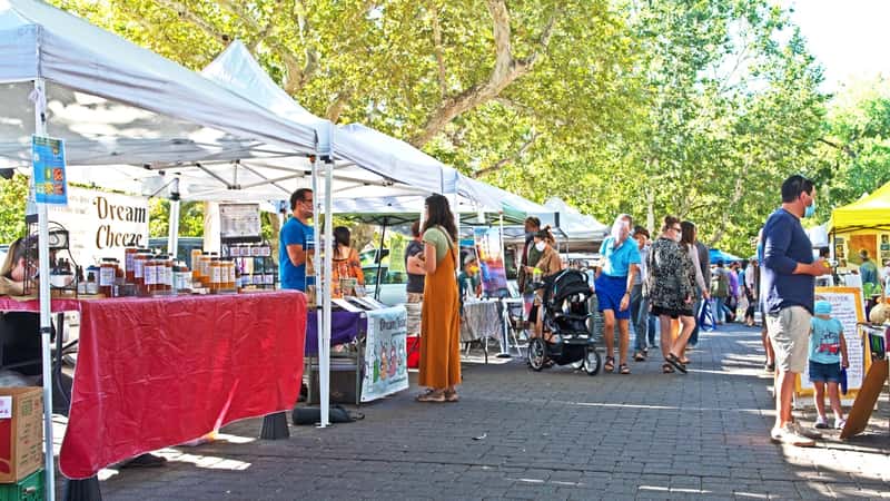 A photograph depicting the Sedona Farmers Market in Sedona, Arizona. The image captures a scene of vendors' stalls, showcasing fresh produce and goods. Visitors can be seen browsing the market, suggesting a typical day of shopping and browsing.