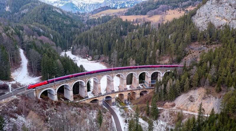 The image depicts an expansive railway bridge, a part of the UNESCO World Heritage Site Semmering Railway in Austria. A lengthy red train, comprising numerous carriages, travels along the tracks spanning the entire length of the bridge. The surroundings feature a vast territory with mountains, hills, and forests, emphasizing the scenic landscape surrounding the railway infrastructure.