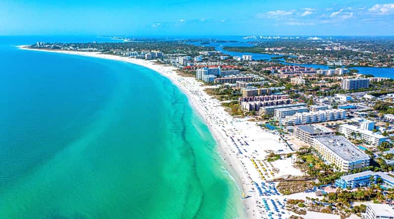 Siesta Beach in Florida is a stunning destination that features powdery, white-quartz sand, crystal-clear waters ideal for swimming, and family-friendly activities like beach volleyball, paddleboarding, and kayaking. Aerial view of blue-green water, long white sand coastline, and many beautiful hotels and buildings. The beach is equipped with umbrellas and sun loungers.