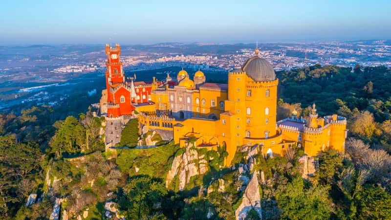 The image features the National Palace of Pena near Sintra, Portugal. The palace stands out with its vibrant colors and unique architecture set against a backdrop of lush greenery. The surrounding landscape includes rolling hills and forests.