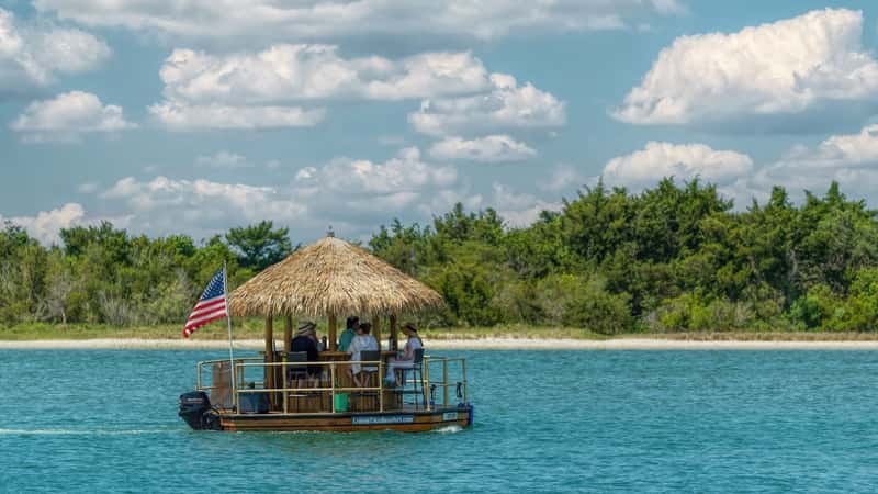 The image features a Tiki Boat Cruise in St. Augustine, showing a boat with a roof made of yellow hay floating next to the USA flag. The scene captures a leisurely boating experience with passengers on board enjoying the outing.