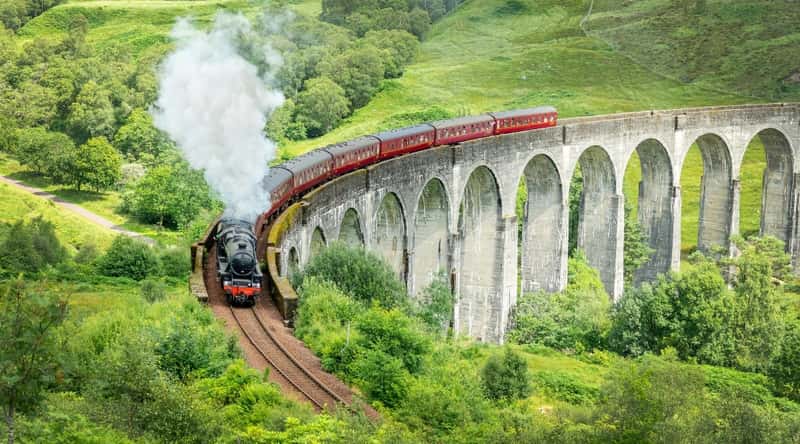 An aerial view capturing the scenic landscape as The Jacobite Steam Train traverses a picturesque bridge. The train, comprising six carriages, emits white steam, creating a classic visual element. The surroundings feature abundant greenery.