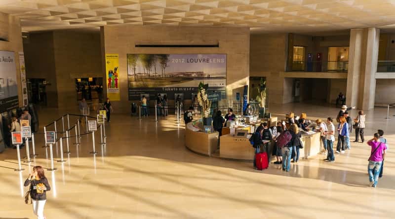 An interior photograph of the Louvre Museum ticket office in Paris, France. The image captures the functional aspect of the ticket office with patrons engaged in purchasing tickets.