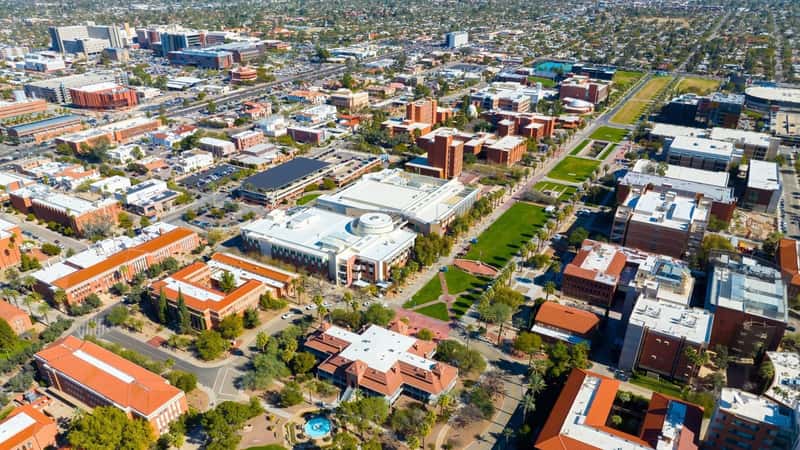 The aerial view of the University of Arizona's main campus in Tucson, Arizona, includes University Mall and the Old Main Building. The surrounding city of Tucson is visible, highlighting a mix of urban and residential areas.