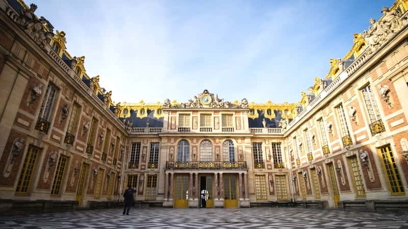 The image shows the front facade of the Chateau de Versailles in Paris, France. The building's architectural style reflects classical French design, featuring symmetrical elements and ornate detailing. The focus is on the exterior of the chateau, emphasizing its grandeur and historical significance.