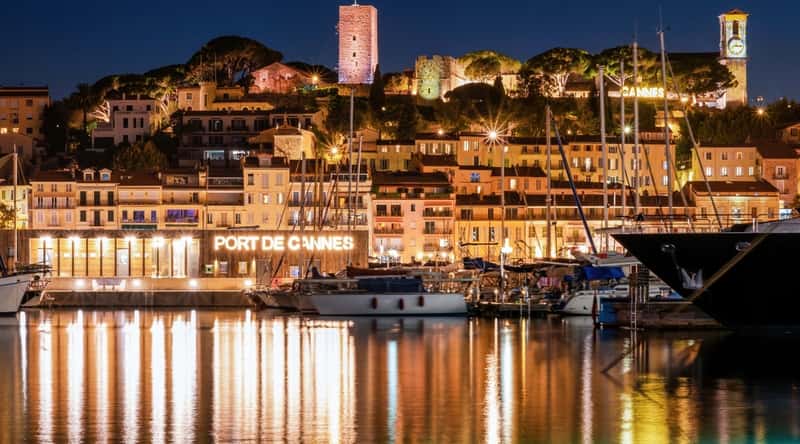 The Vieux Port in Cannes France shot from the waterfront at night with the city lit up and the reflection shimmering off the water.