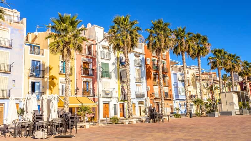 The image portrays Villajoyosa, a scenic coastal town located in southern Spain. The town features low buildings, with houses arranged in a row, each painted in vibrant colors, adding to the picturesque charm of the area. In front of the houses, there are palm trees that are as tall as the buildings, contributing to the coastal ambiance. The foreground of the image shows sandy terrain, typical of coastal regions.
