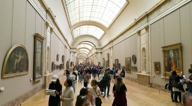 A depiction of visitors walking through a long, well-lit room within the Louvre museum in Paris. The space is adorned with paintings along the walls, illuminated by natural light from a glass ceiling.