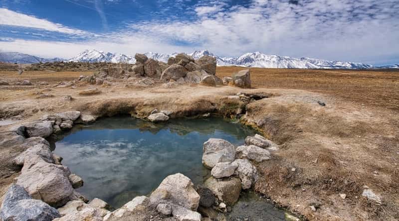 A photograph depicting the landscape around Wild Willy's Hot Springs in California. The image captures the natural hot springs within a desert environment. The pools are visible amidst the arid terrain, surrounded by a minimalistic setting with no prominent architectural features.