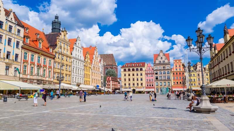 The image captures the city center and Market Square in Wroclaw, Poland. The square appears relatively empty, with few people present. Surrounding the square are rows of brightly colored row houses. The architecture reflects a mix of historical and modern elements typical of Wroclaw's cityscape.